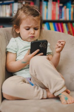 Kanepede oturan ve cep telefonuyla sosyal medyada gezinen gündelik kıyafetlerle tatlı küçük bir kız.