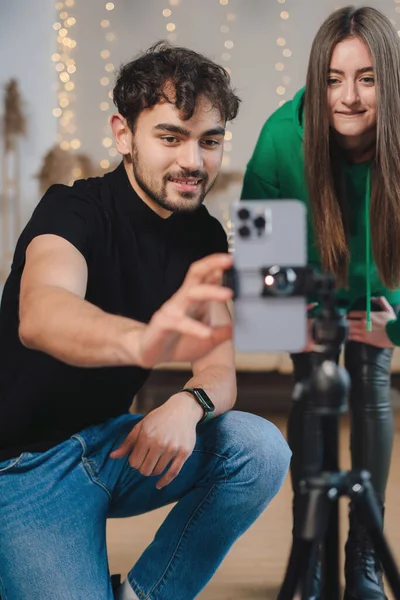 Neşeli genç adam ve kadın cep telefonuyla selfie çekerken rahat bir odada Noel için süslenmiş bir şekilde oturuyorlar.