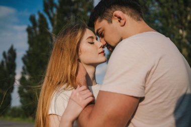 Yeşil ormanda dikilen genç bir adam ve kadının birbirine sarılıp öpüşmesi.