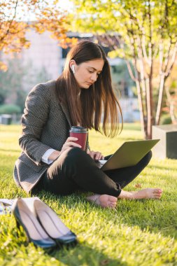 Güneşli bir günde parkta dinlenirken çimde oturan ve tablet okuyan gündelik kıyafetli genç bir kadın.