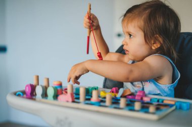 Aydınlık odada masada oturmuş renkli oyuncaklarla oynayan, günlük kıyafetlerle gezen küçük tatlı bir kız.