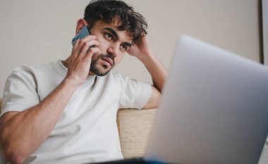 Gündelik giysiler içinde genç bir erkek dizüstü bilgisayarla masada oturuyor ve evde uzak bir proje üzerinde çalışırken cep telefonuyla konuşuyor.