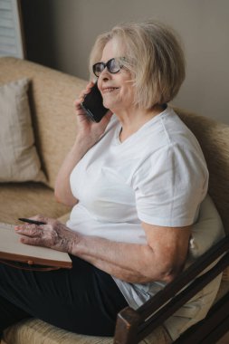 Normal giyinen ve gözlüklü yetişkin bir erkeğin ahşap masada oturması ve evdeki rahat oturma odasında dinlenirken akıllı telefondan fotoğraf çekmesi.