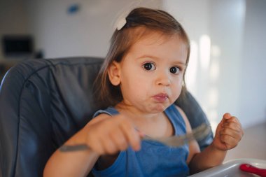 Evdeki aydınlık oturma odasındaki kanepede oturmuş kameraya bakan gündelik giysiler içindeki sevimli küçük kız.