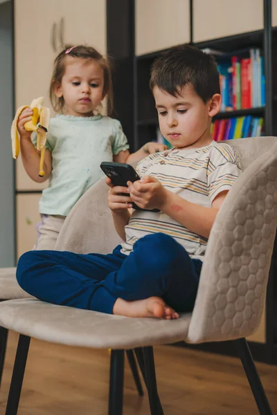 穿着休闲装的可爱小男孩一边坐在舒适的客厅桌子旁一边浏览智能手机 一边看着姐姐在吃香蕉 — 图库照片