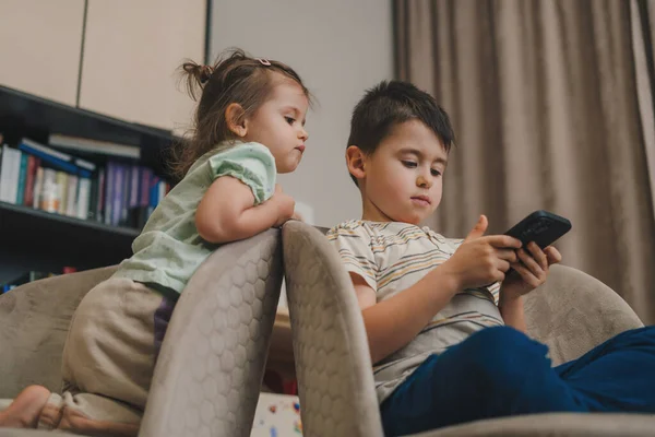 可爱的小男孩坐在沙发上玩电子游戏 而她的姐姐却看着他呆在家里 — 图库照片