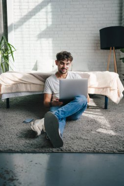 Evde uzak bir proje üzerinde çalışırken kanepede oturup laptopunu karıştıran, günlük kıyafetlerle serbest çalışan sakallı bir erkek.