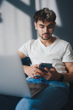Ciddi bir genç adam, gündelik giysiler içinde kanepede oturuyor ve evde uzaktan kumandalı bir proje üzerinde çalışırken akıllı telefonlara bakıyor.