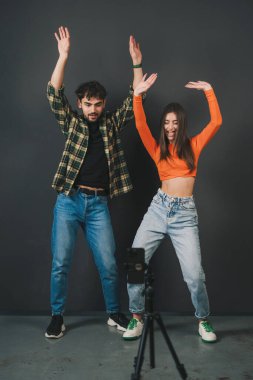 Stüdyoda siyah arka planda dikilirken kollarını kaldırıp kameraya bakan modaya uygun giyinmiş genç bir kadın.