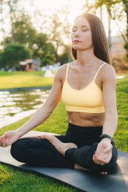 Spor kıyafetli genç bir kadın vücudu parkta yoga yaparken mindere oturup bacaklarını esnetiyor.