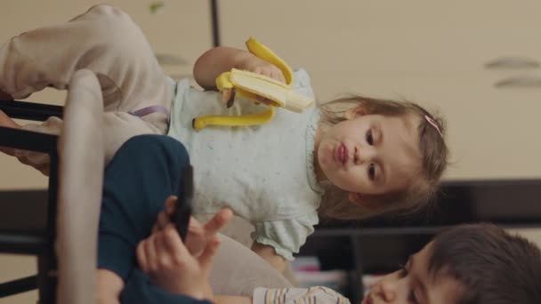 Egy Kislány Banánt Eszik Étvággyal Nézi Ahogy Bátyja Mobilon Játszik — Stock videók