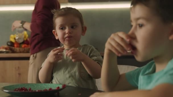 キッチンテーブルに座っているかわいい陽気な少年と赤ん坊の少女は 彼らの目の前にジューシーな赤い新鮮なザクロの種子でいっぱいのプレートです 健康的に食べる ライフスタイル — ストック動画