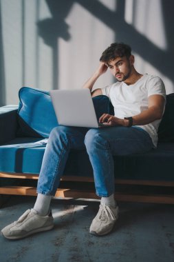 Kafkasyalı bir adam dizüstü bilgisayarda çalışıyor, oturma odasındaki koltukta oturuyor. Mesafe işi, serbest çalışma, sosyal ağlarda iletişim.