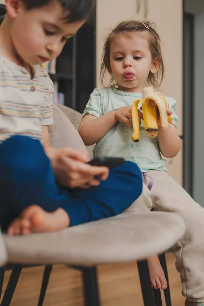 Hermanita Comiendo Plátano Mirando Hermano Jugando Teléfono Móvil Gadget Adictos — Foto de Stock