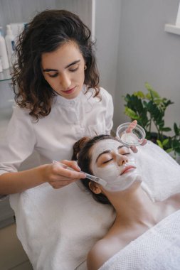 Kozmetik uzmanı kadın müşteriye yüz maskesi takıyor, spa salonunda işlem yapıyor. Estetik kliniği. Cilt bakımı, kozmetoloji ve güzellik.
