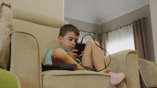 白人小男孩坐在沙发上玩着一个手里拿着便携式游戏机的游戏 现代游戏设计 流行的游戏 玩孩子 — 图库视频影像