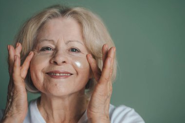 Gri saçlı güzel bir kadının portresi cilt bakımını yapıyor, göz bandı takıyor. Stüdyo çekimi, hafif ışıklandırma. Makyaj yok, cildi temiz.