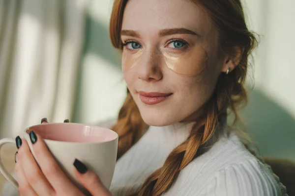Portrait of smiling attractive woman looking at camera with eye patches holding a cup of coffee in the living room. People lifestyle portrait. Domestic
