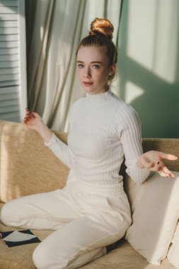 Genç, çekici, esmer kızıl kanepede bağdaş kurmuş oturuyor. Kameraya bakıyor ve el kol hareketi yapıyor. İnsanların yaşam tarzı portresi. Sağlıklı yaşam tarzı, sağlık.