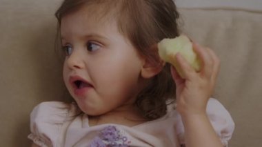 Evdeki kanepede oturmuş elma yiyen küçük bir kız. Sağlıklı beslenme. Sağlıklı yiyecekler, diyet. Organik yiyecekler. Lezzetli yemek..