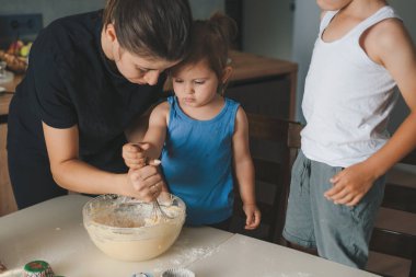 İki küçük çocuk anneleriyle birlikte mutfakta kek yapmak için hamur işi hazırlıyorlar. Çocuklar için aile aktiviteleri.