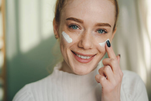 Рыжая молодая женщина наносит увлажняющий крем на лицо веснушками для здоровой кожи вокруг глаз. Реклама косметики Skincare