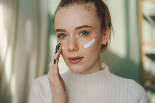 Обрезать молодую женщину с короткими волосами наносить крем на лицо и смотреть в камеру, стоя в светлом помещении