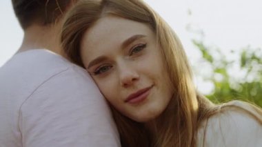 Bahçedeki beyaz bir çiftin yakın görüntüsü. Kameraya bakıp gülümsüyorlar. Kızıl saçlı kadın, başını erkeğin omzuna yaslıyor. Güzel aile..