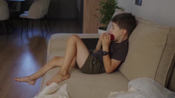Αγόρι Δαγκώνει Κόκκινο Μήλο Ενώ Βλέποντας Κινούμενα Σχέδια Στην Τηλεόραση — Αρχείο Βίντεο
