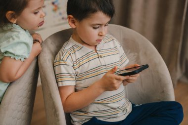 İki çocuk telefon bağımlısı, telefon kullanıyorlar, koltuklarda oturuyorlar. Akıllı telefon bağımlılığı konsepti. Güzel bir aile. Çocuğu oynuyorum. Çevrimiçi oyna.