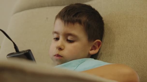 ティーンエイジャーの少年はソファーに横たわり 彼の手にポータブルゲームコンソールを持っているゲームをプレイします 10代はどのように休暇を過ごすか — ストック動画