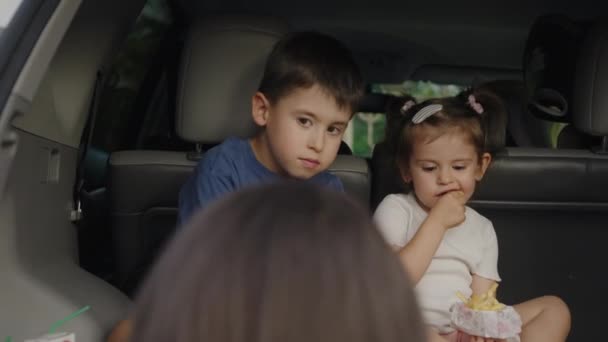 ハッピーキューティー 小さな女の子と少年は 夏の旅行中に車のトランクに座っているおいしいスナックを持っています 若い家族夏休みや休日 — ストック動画