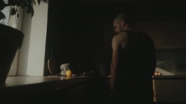 Siyah tişörtlü bir adamın mutfakta dolaşıp o sabah için ne hazırlayacağını düşünmesi. Sabahları mutfağın karanlık manzarası.