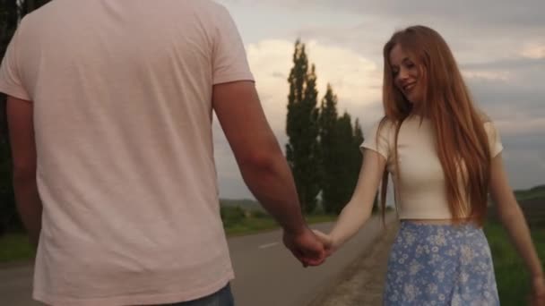 年轻的高加索红头发女人 穿着蓝色的裙子 牵着男朋友的手 走在路边 美丽的夫妻在外面消磨时光 欣赏乡村美景 — 图库视频影像