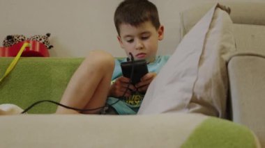 Küçük beyaz çocuk kanepede oturmuş, el bilgisayarı oyunu oynuyor. Neşeli çocuklar video oyunu oynuyor