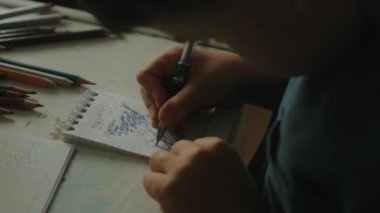 Küçük bir çocuğun yazı yazarken, defterine çizim yaparken, masada otururken ve ödev yaparken yakın plan görüntüsü. Evde eğitim ve eğitim gördüm. Okula geri dön..