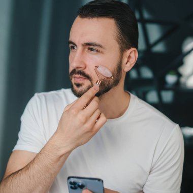 Yüzüne parlak cilt masajı yapan yeşim taşlı silindir kullanan bir adam. Yakışıklı genç adam yaşlanmayı önleyen bir güzellik taşıyla pratik yapıyor..