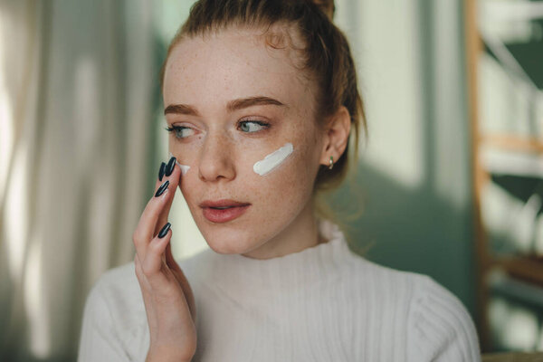 Привлекательная женщина с веснушками, лежащая на диване и трогающая лицо пальцами с помощью ежедневного увлажняющего крема дома. Красота и скинхед