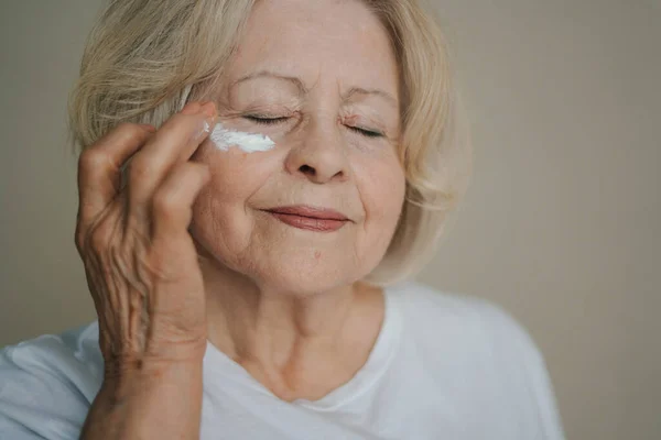 Lovely senior woman applying face cream under her eyes over studio background. Older lady taking care of her skin. Mature beauty concept.
