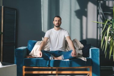 Yumuşak bir kanepede rahat bir şekilde oturan, bacakları çapraz ve gözleri meditasyon yaparken kapalı rahat bir adamın tam boy görüntüsü. Uyum sağlayan temel yoga egzersizi