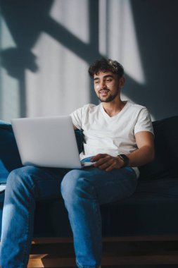 Kanepede oturan genç adam dizüstü bilgisayarını kucağında tutuyor ve internette geziniyor. Sosyal ağlar. E-alışveriş.