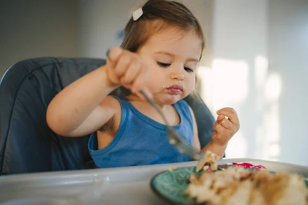 Adorable baby girl eating with an appetite holding fork sitting in highchair and learning to eat by itself. Food, child, feeding and development concept.