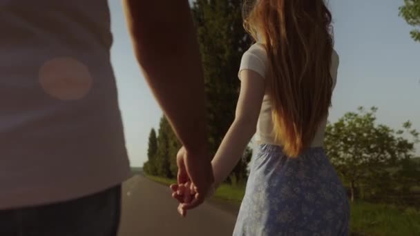 一个男人和红头发的女人在美丽温暖的日落中走在路上的背影 女人牵着男人的手 — 图库视频影像
