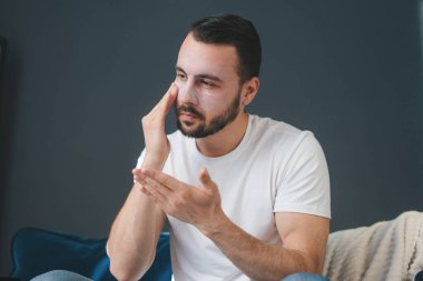 Yakışıklı bir adam evdeki kanepede otururken yüzüne nemlendirici krem sürüyor. Parmaklarıyla tenine dokunuyor. Spa modeli, nemlendirici krem.
