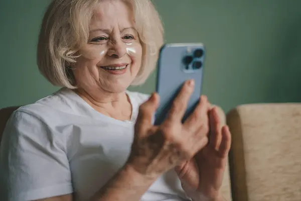 Elderly woman in a white t-shirt texting and watching video on the phone while sitting on the sofa in the evening and relaxing. Video call, new app, online