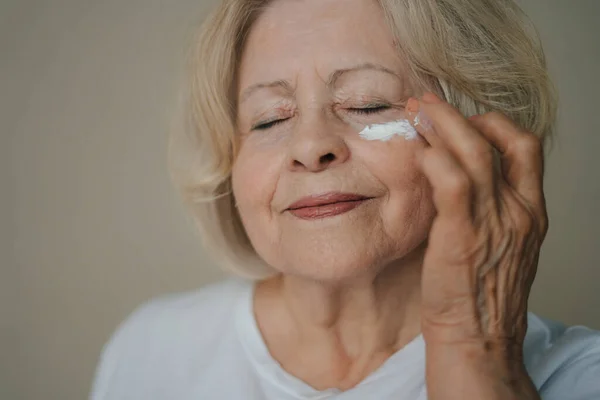 Close-up portrait of beautiful senior woman gently applying under eye face cream while closing her eyes. Product for beauty skincare