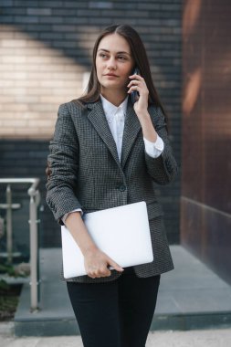 Genç, kumral bir kadın elinde dizüstü bilgisayarla telefonda konuşuyor. Çekici bir kadın şık siyah ceket giyiyor, başka tarafa bakıyor. Çevrim içi iş