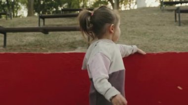 Beyaz küçük bir kız elinde kırmızı bir duvara dokunarak parkta yürüyor. Çocukluk hayalleri. Aile, doğa, özgürlük. Sevimli küçük çocuk..
