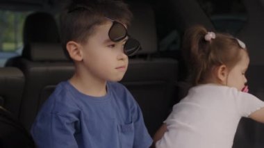 Komik çocuk arabada kız kardeşiyle oturuyor, yüzünde güneş gözlüğü var ve eğri büğrü duruyor. Çocuk, doğada ılık yaz gününün tadını çıkarıyor.