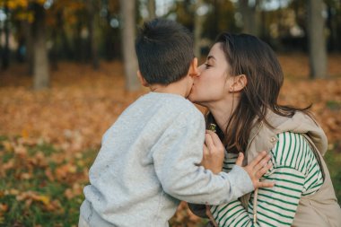 Küçük oğlunu yanağından öpen mutlu genç bir kadın sonbahar parkında eğleniyor. Anne ve oğlu parkta yürüyor. Aşk ve aile kavramı.